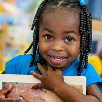 A smiling pre-school student clutches a book to her chest at the Early Steps for School Success program (ESSS) in Barnwell, South Carolina: Photo credit: Susan Warner/Save the Children, October 2014. A smiling pre-school student clutches a book to her chest at the Early Steps for School Success program (ESSS) in Barnwell, South Carolina: Photo credit: Susan Warner/Save the Children, October 2014.
